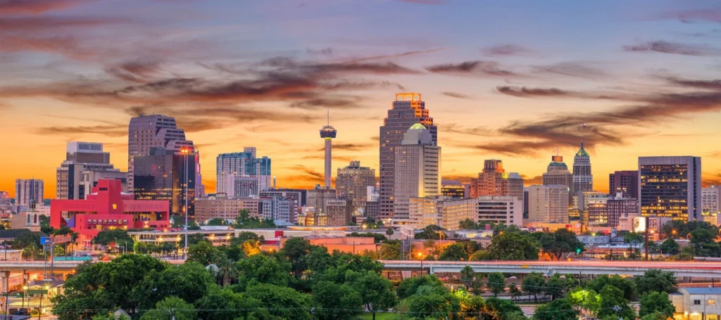 San Antonio city skyline at sunset, with tall buildings, the Tower Life Building, and the Tower of the Americas visible against a colorful sky with orange, yellow, and blue hues. Trees and highways are seen in the foreground.