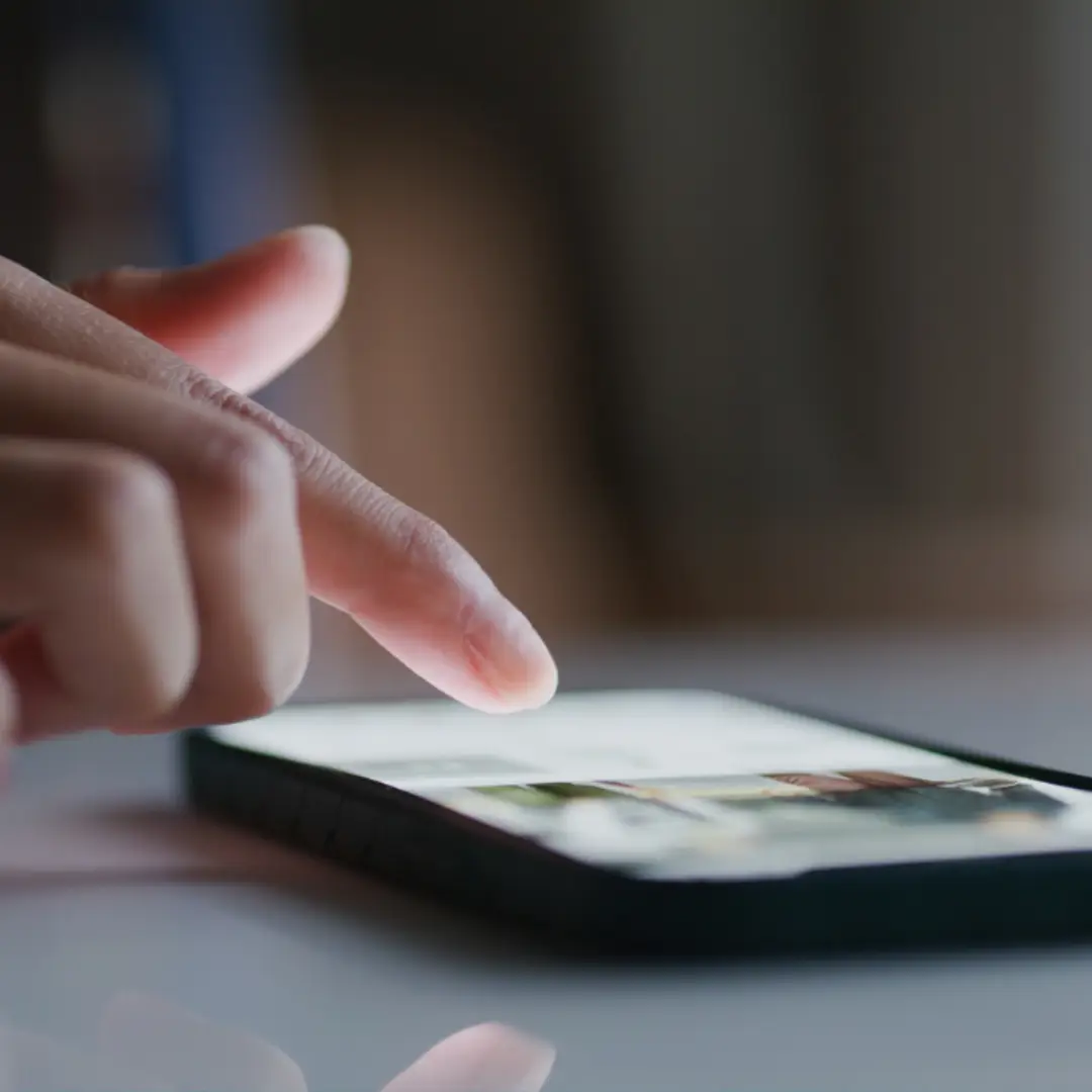 A close-up of a persons hand swiping or tapping on a smartphone screen, with the device resting on a reflective surface in a dimly lit environment.
