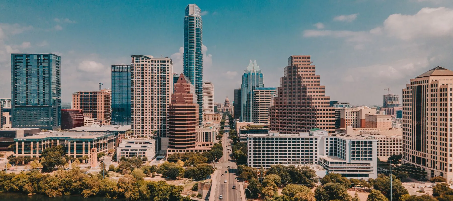 Aerial view of downtown Austin, Texas, featuring modern high-rise buildings, a clear blue sky, and a busy street lined with trees in the foreground.
