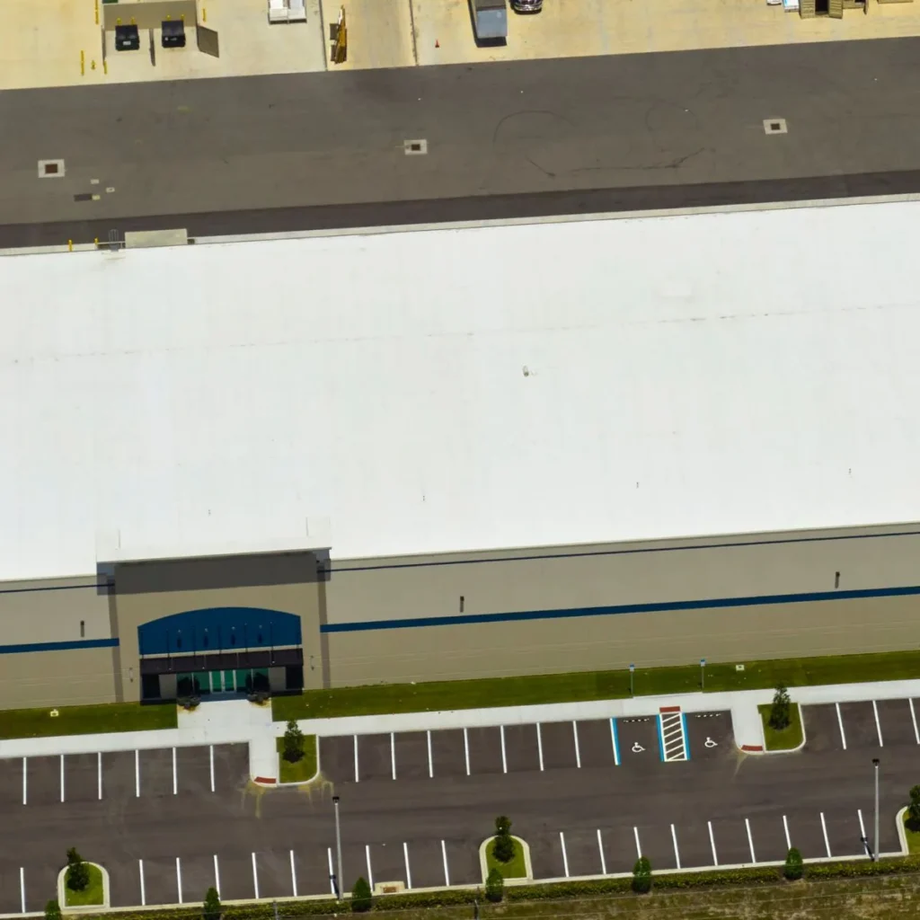 Aerial view of a large warehouse building with a white roof, an empty parking lot in front, and several designated accessible parking spaces near the main entrance.