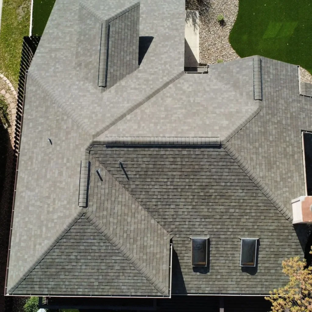 Aerial view of a house roof with gray shingles, multiple ridges, dormers, and vents; surrounding areas include green lawn, gravel, and trees.