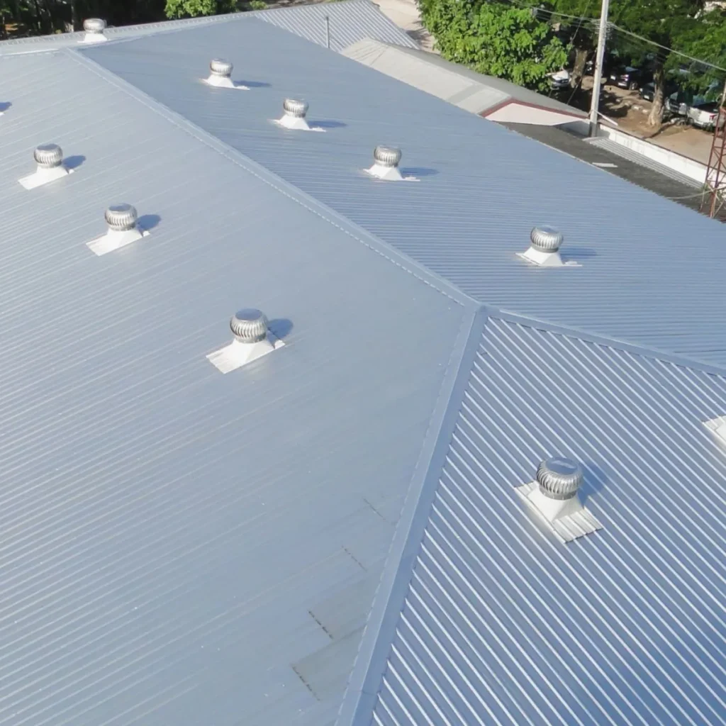 Aerial view of a large corrugated metal roof with multiple round roof ventilators evenly spaced across its surface; trees and a few buildings are visible in the background.