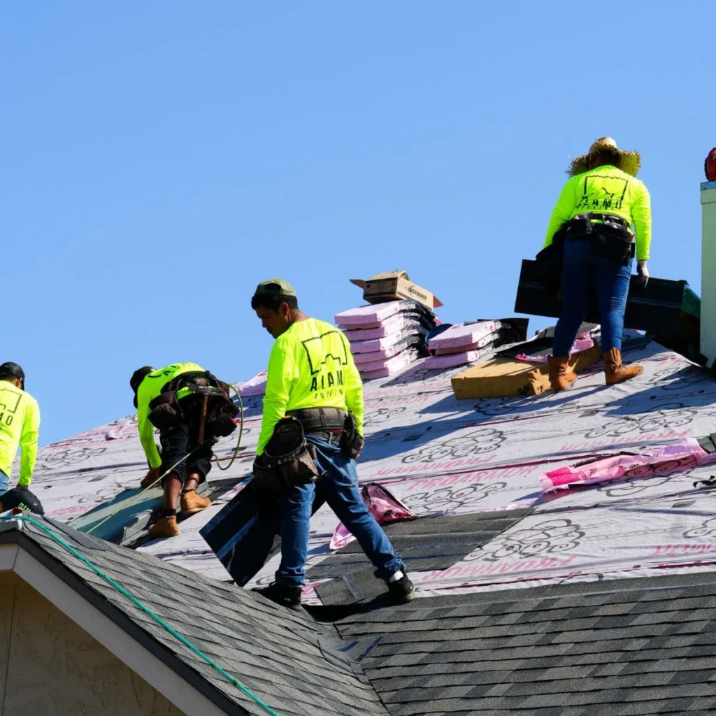 Four workers in bright yellow shirts install roofing materials on a sloped house roof under a clear blue sky. Packs of shingles are stacked on the roof, and safety harnesses are visible.