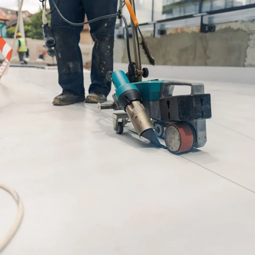 A worker operates a floor welding machine to seal seams on a flat, white surface. The machine emits heat along the seam, while construction materials and a partially built wall are visible in the background.
