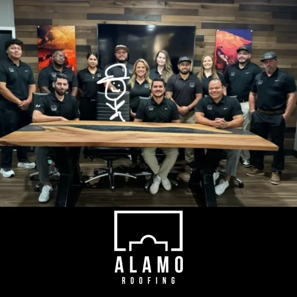 A group of eleven people in matching black shirts pose indoors around a wood table, smiling at the camera. Behind them is a stick figure drawing on a whiteboard. The Alamo Roofing logo appears at the bottom of the image.