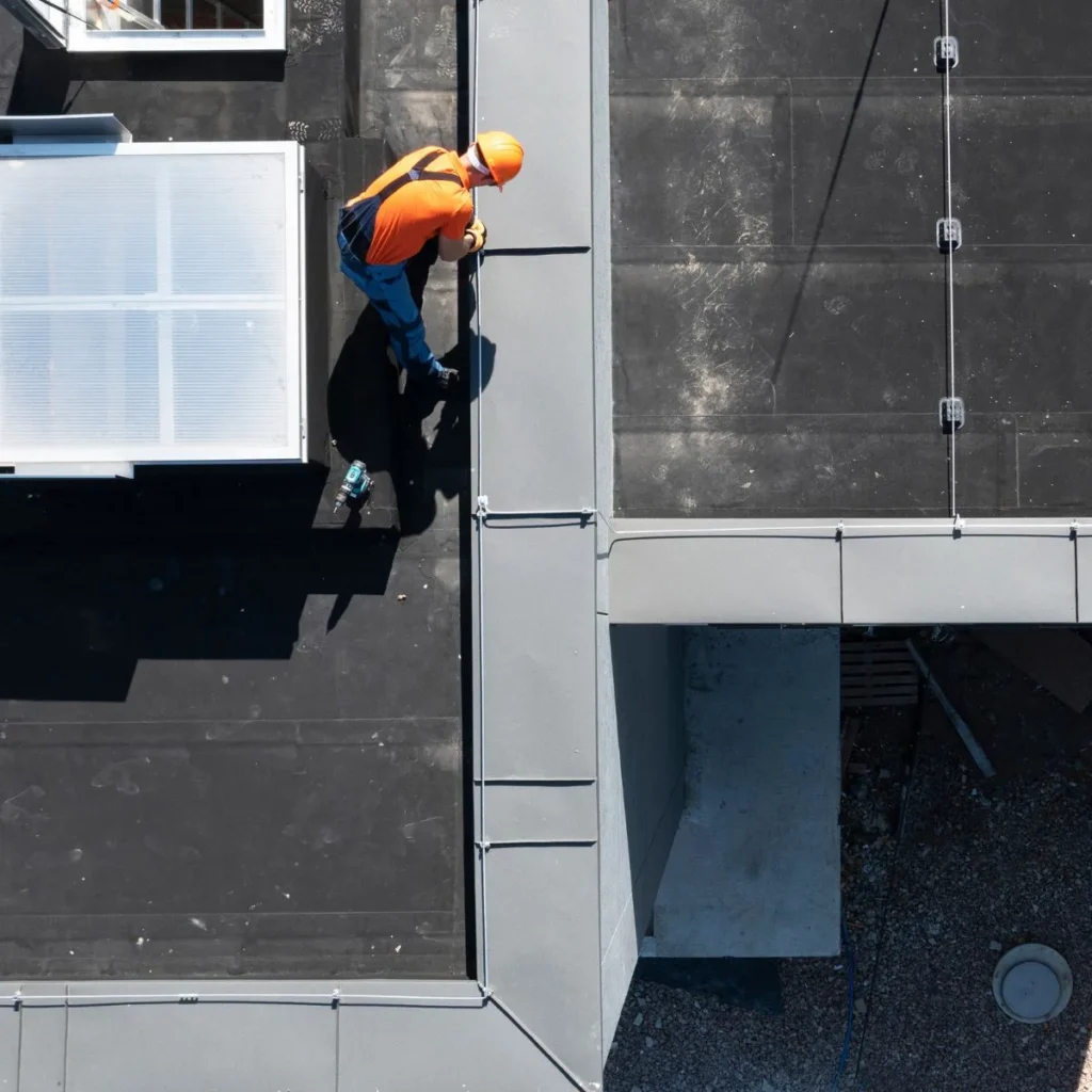 A worker wearing an orange helmet and shirt installs or repairs metal flashing on a flat, black rooftop, viewed from above. Tools are placed nearby, and part of a skylight is visible.