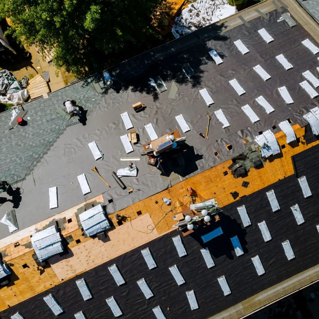 Aerial view of workers installing new shingles and underlayment on a house roof, with tools and materials scattered across the surface. Some sections show exposed wood, while others have black roofing paper.