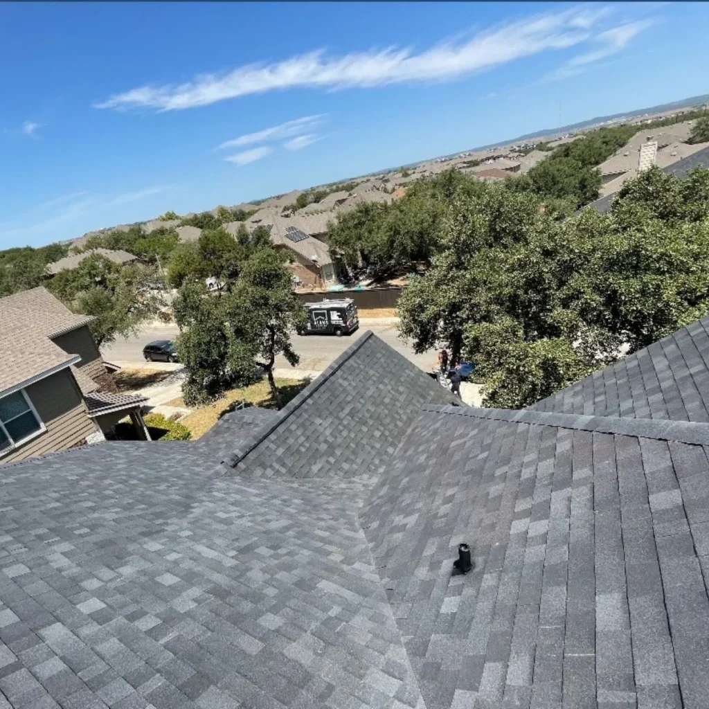 View from a rooftop with gray shingles, overlooking a neighborhood with houses, trees, and a clear blue sky. A truck is parked on the street below.