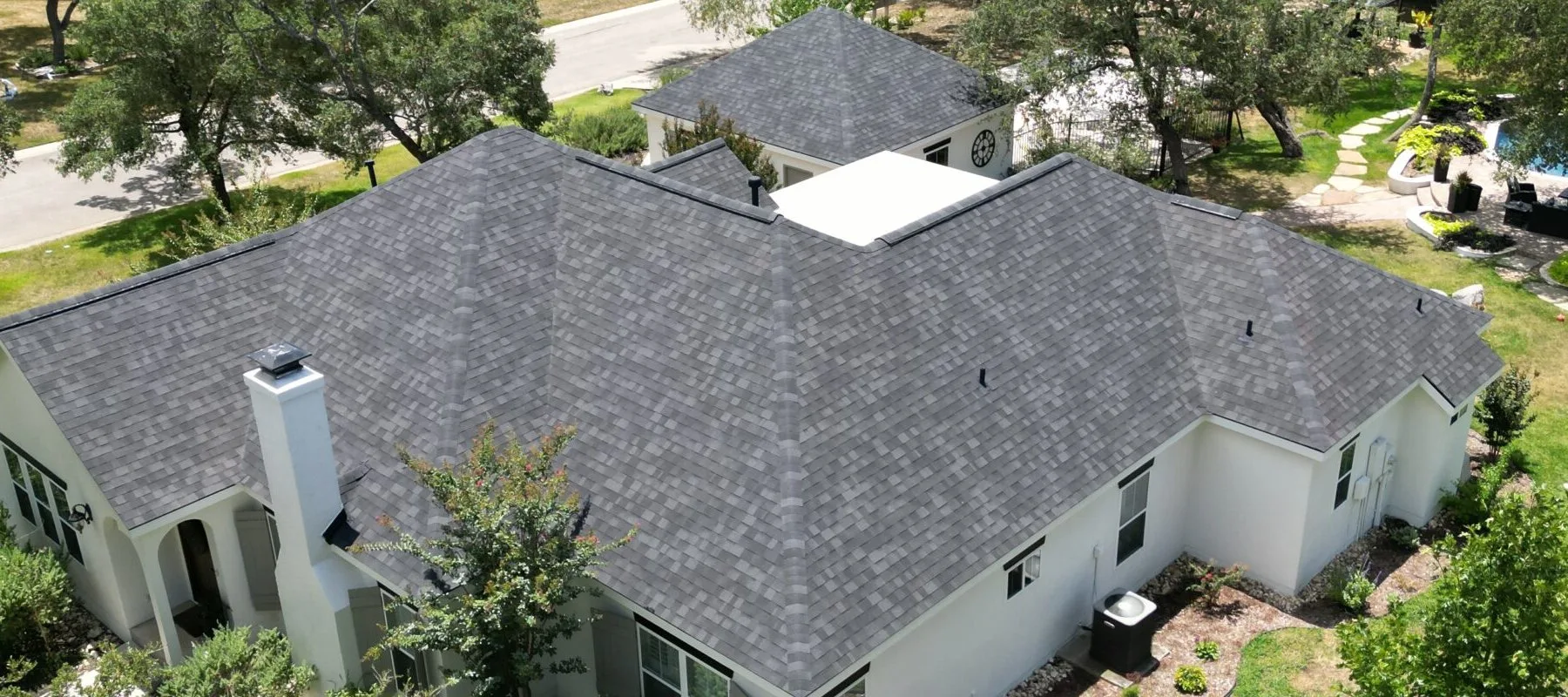 Aerial view of a house with a gray shingle roof, white exterior walls, and surrounding greenery, including trees and shrubs. Nearby, a smaller building with a matching roof is visible.
