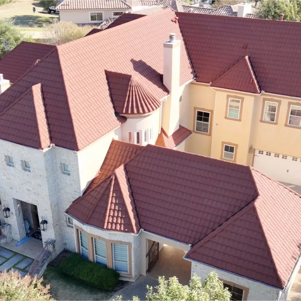 Aerial view of a large, two-story house with light-colored stone and stucco walls, multiple red tile roofs, chimneys, and arched windows. The property is surrounded by trees and neighboring homes.