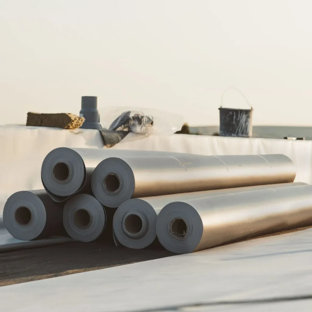 Six rolls of roofing or construction membrane are stacked on a rooftop, with various building materials and a bucket visible in the background under soft daylight.