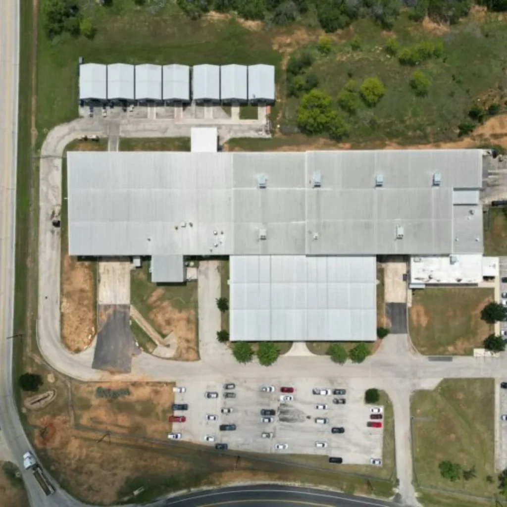 Aerial view of a large industrial or commercial building with a metal roof, surrounded by a parking lot filled with cars and some grassy areas with scattered trees. Several smaller structures are visible nearby.