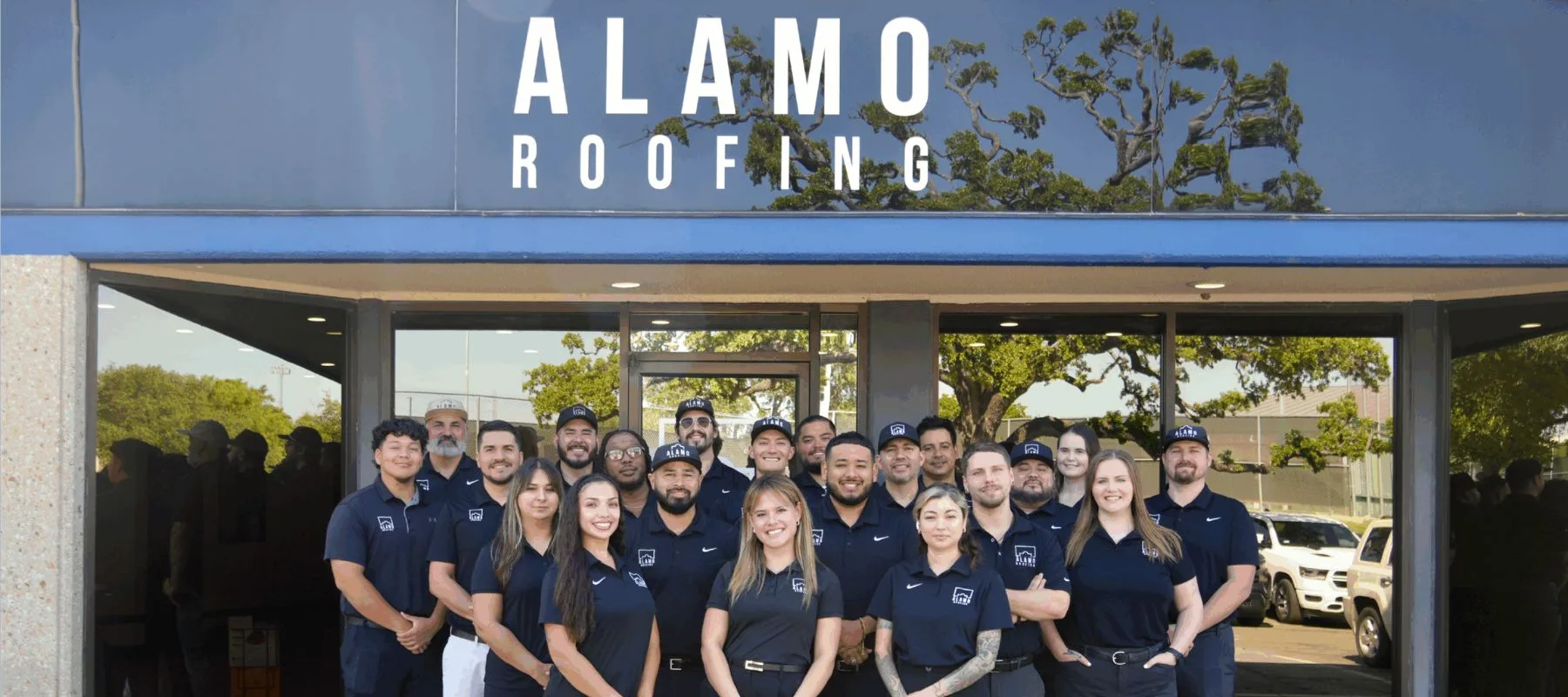 A group of employees wearing matching navy shirts poses and smiles in front of the Alamo Roofing office building, with the company name visible above them in large white letters.
