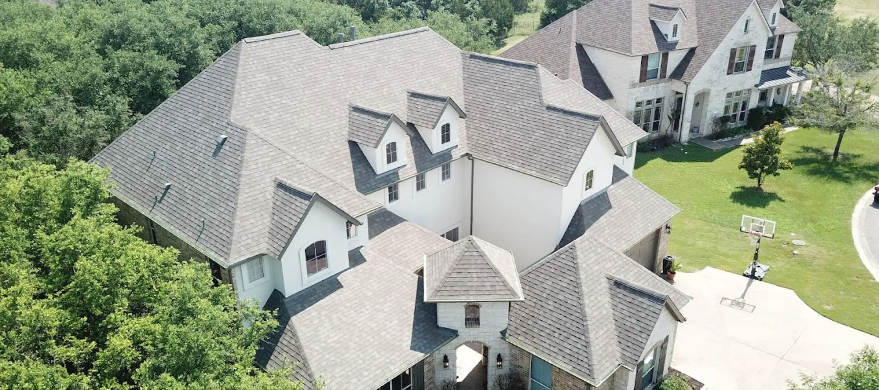 Aerial view of a large, multi-story house with a complex gray shingle roof, surrounded by trees. A driveway with a basketball hoop and another house are visible nearby.