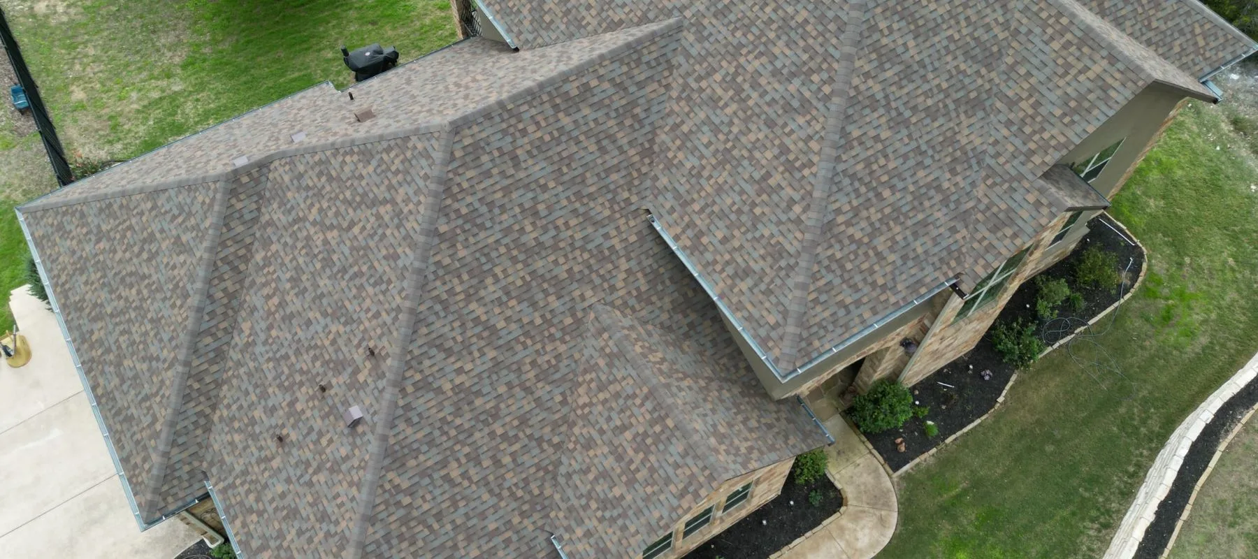 Aerial view of a house with a multi-layered, brown shingled roof, surrounded by green grass, a concrete walkway, and landscaped garden beds.