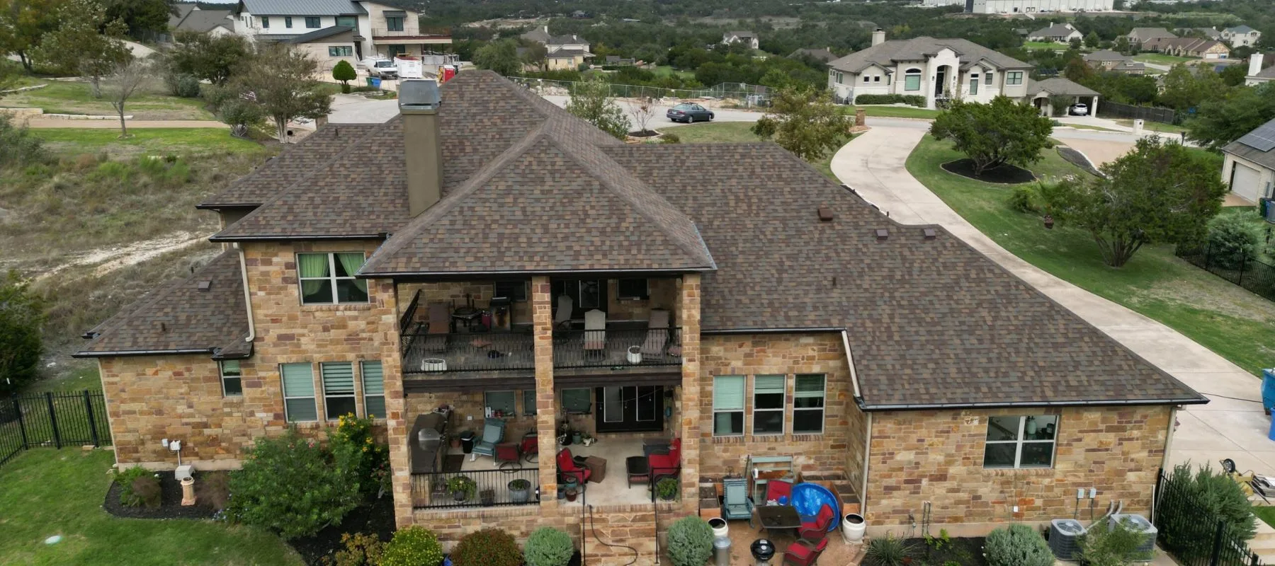A large two-story house with a brown shingle roof and stone exterior, featuring a covered patio, outdoor furniture, and a spacious yard with landscaping in a suburban neighborhood.
