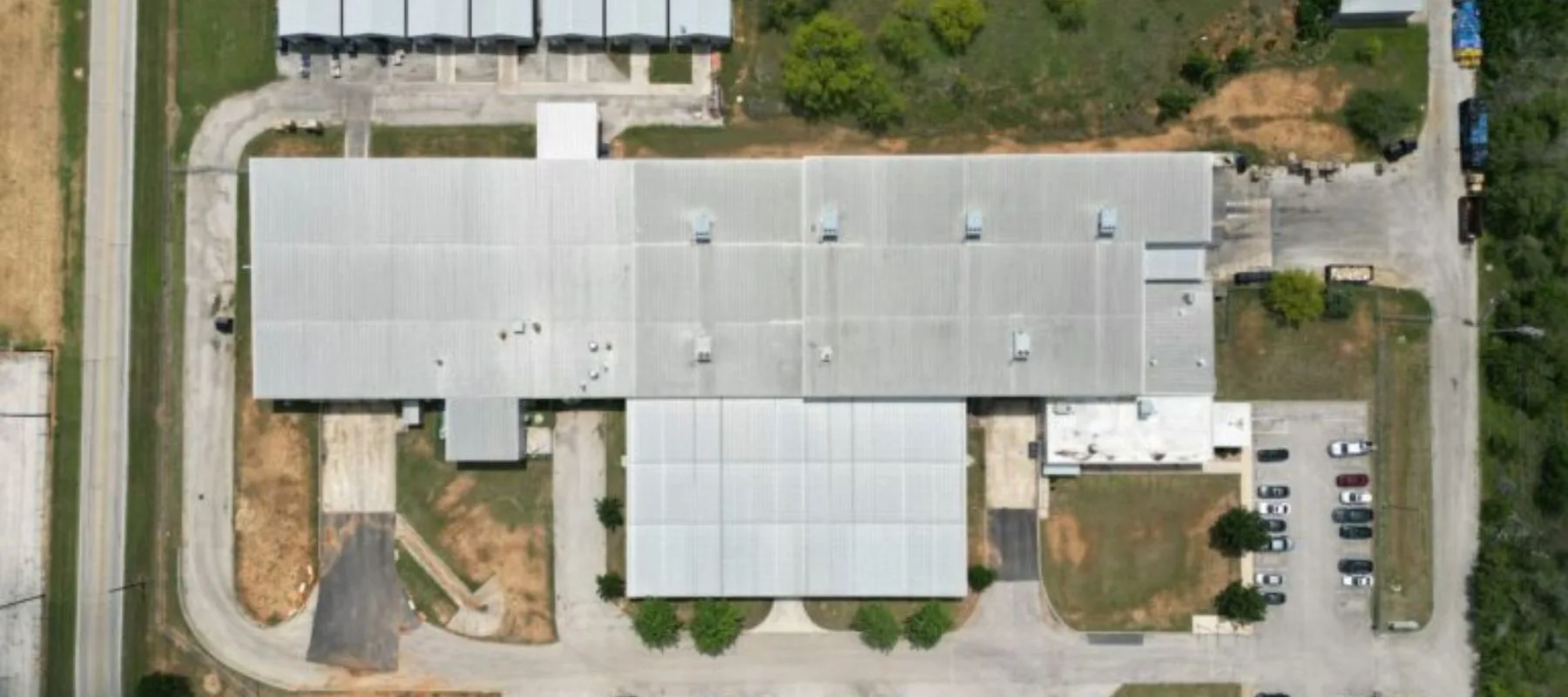 Aerial view of a large, rectangular industrial building with a metal roof, surrounded by parking lots, several cars, access roads, and patches of grass and trees.