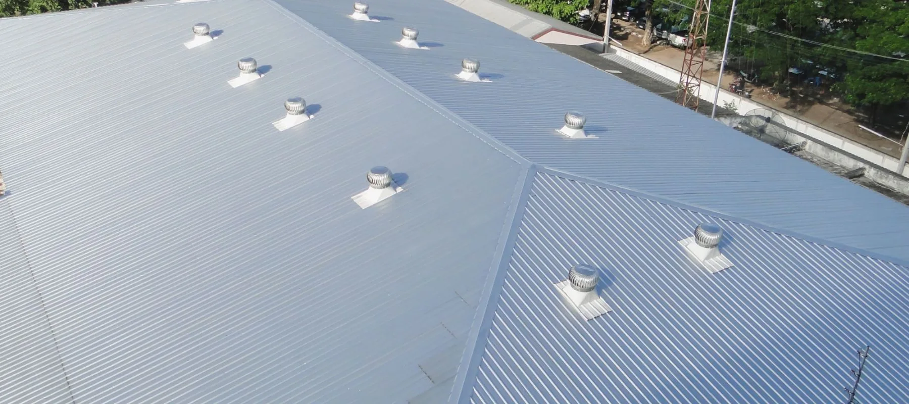 Aerial view of a large, light gray corrugated metal roof with multiple cylindrical roof vents evenly spaced across its surface. Trees and a street are visible in the background.