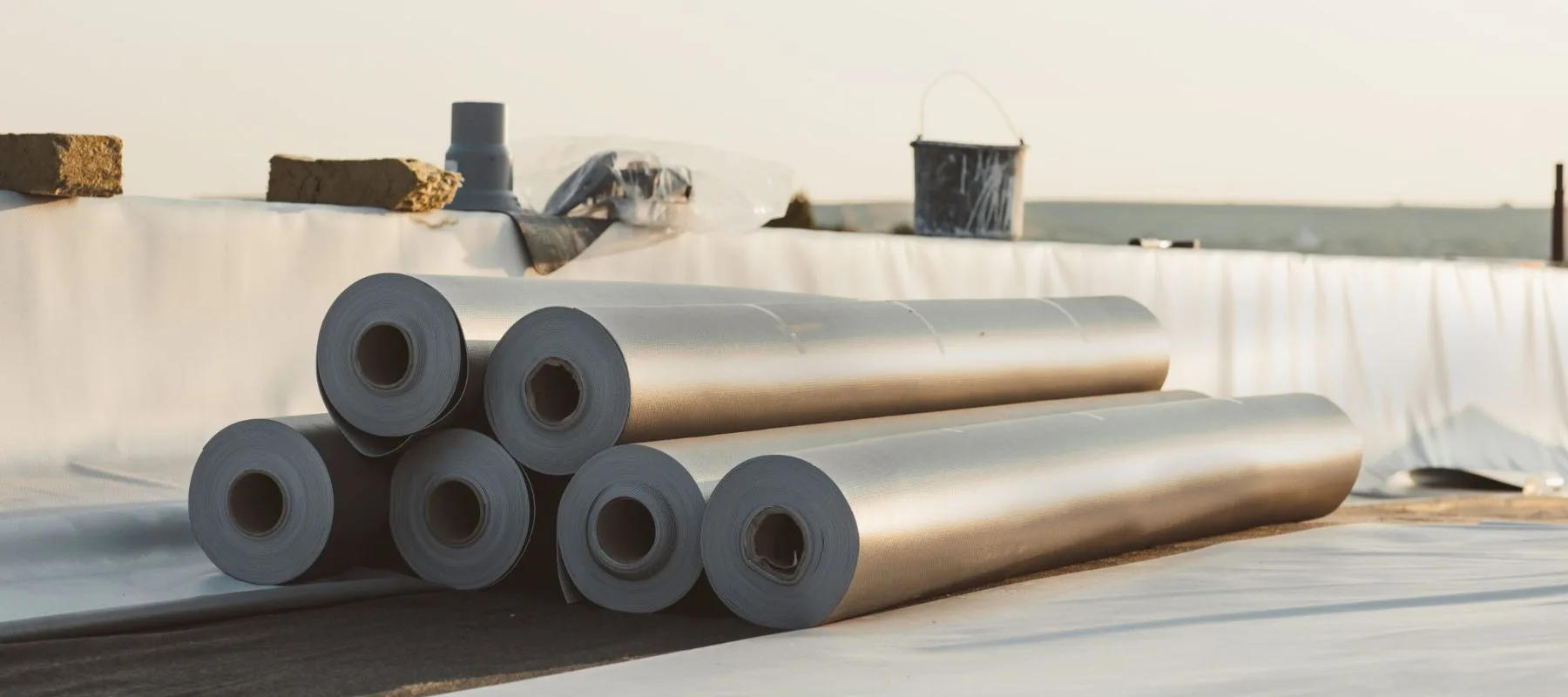Six large rolls of roofing material are stacked on a flat rooftop, with construction tools and supplies visible in the background under soft daylight.