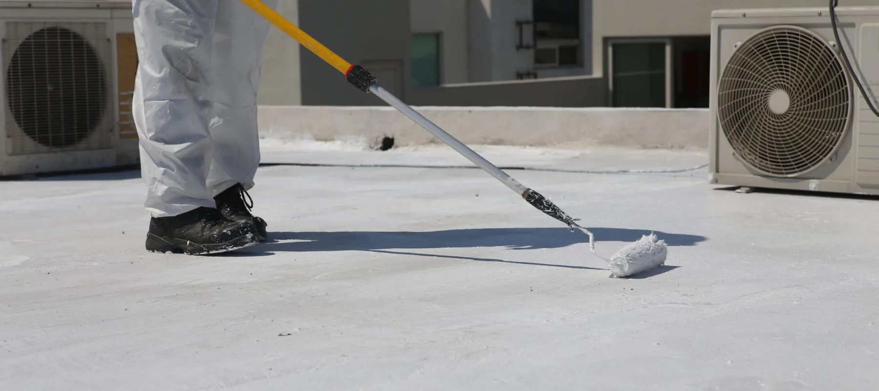 A person wearing protective clothing uses a paint roller on a pole to apply white coating to a flat rooftop, with air conditioning units visible nearby.