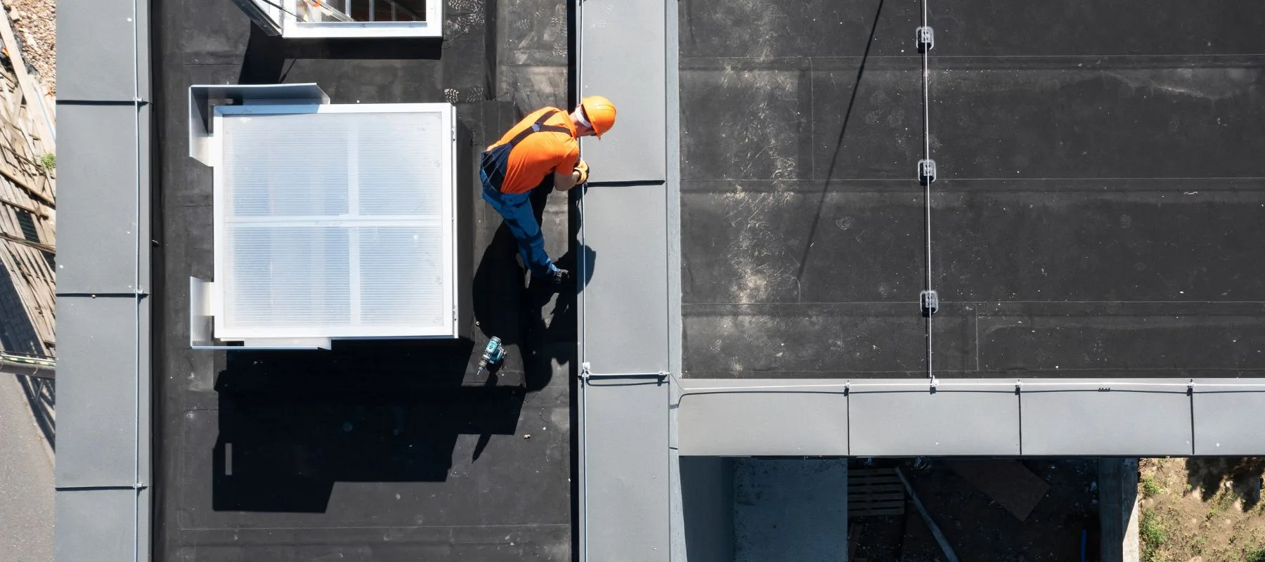 A construction worker in an orange shirt and helmet stands on a flat rooftop, working near ventilation equipment and casting a shadow on the black surface.