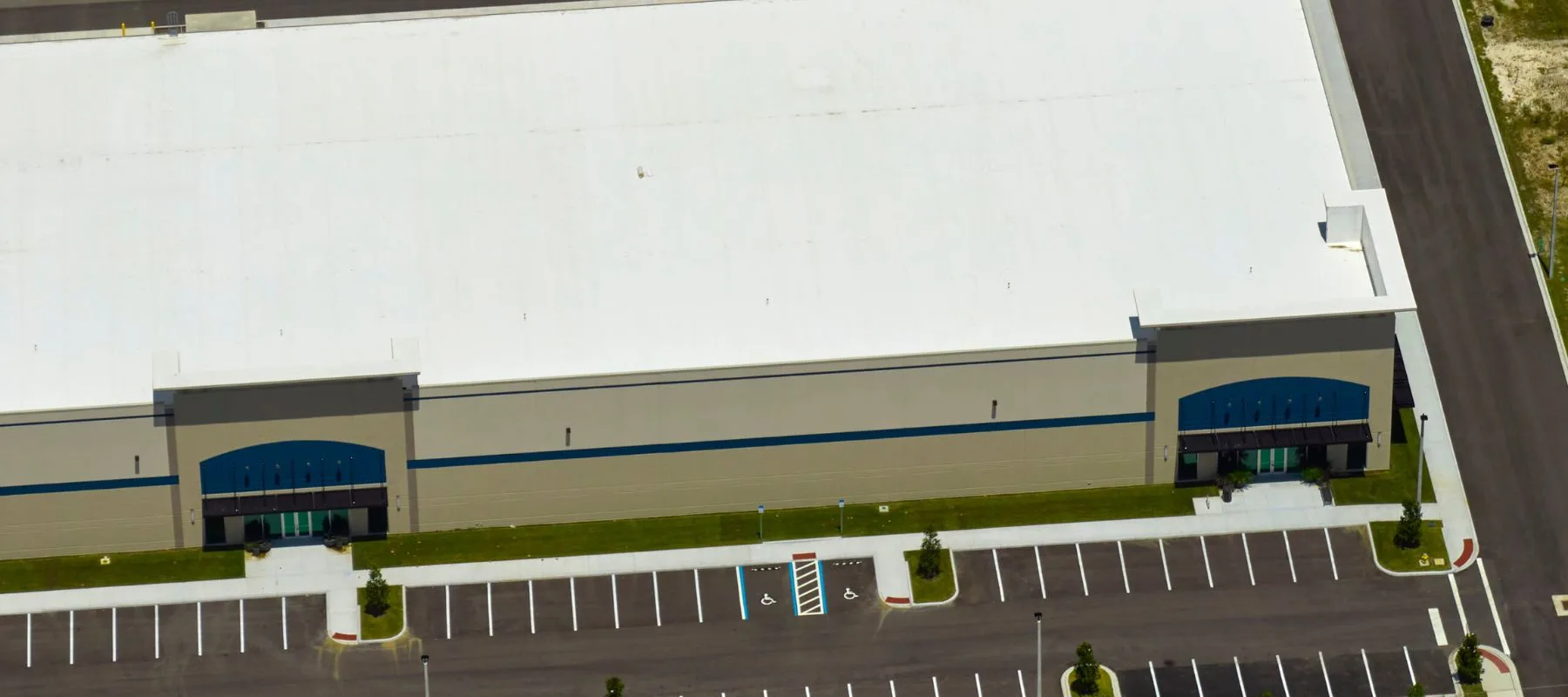 Aerial view of a large, beige industrial building with a white roof, multiple entrances, and an adjacent empty parking lot with marked accessible parking spaces.