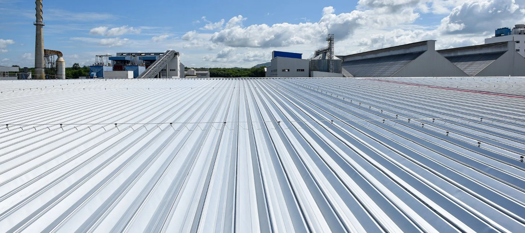 A wide view of a white metal sheet roof on an industrial building, with factory structures and a blue sky with scattered clouds in the background.