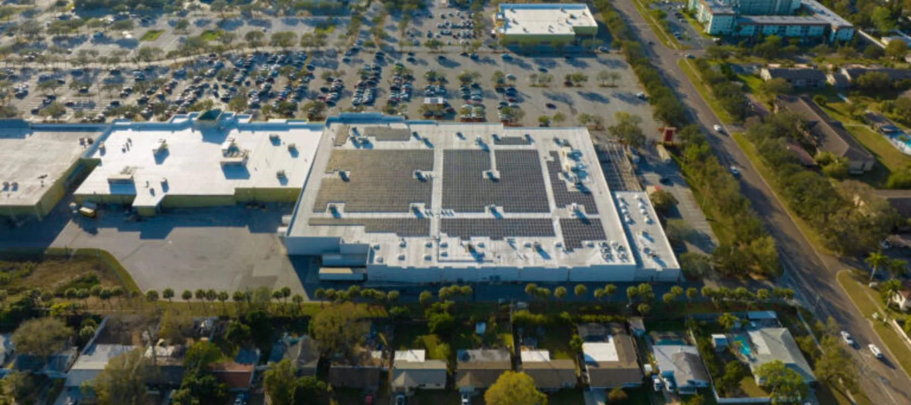 Aerial view of a large commercial building with a white roof and solar panels, surrounded by a parking lot filled with cars, trees, and nearby roads and residential houses.