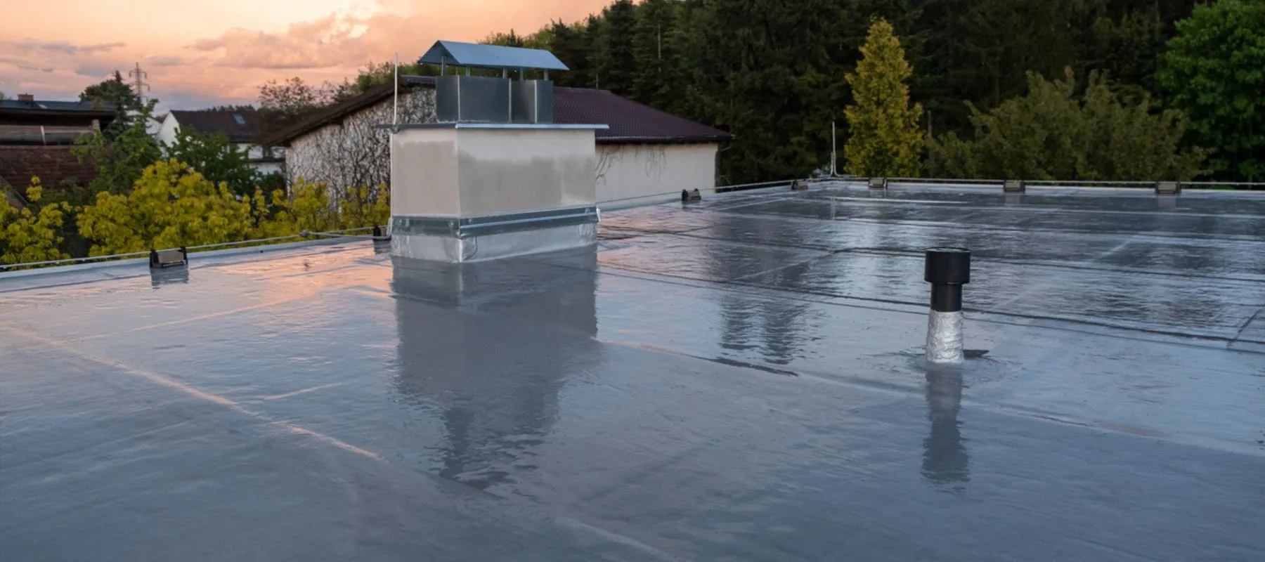A flat rooftop covered with a smooth, reflective waterproof membrane. A small ventilation structure and two vents are visible, with trees and houses in the background during sunset.