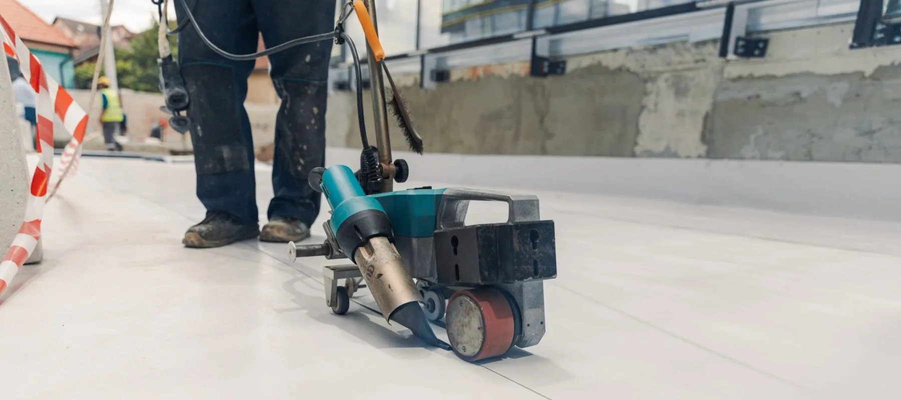 A worker uses a specialized welding machine to fuse seams on a flat, white surface, likely installing a waterproof membrane at a construction site. The worker’s legs and equipment are visible in the foreground.