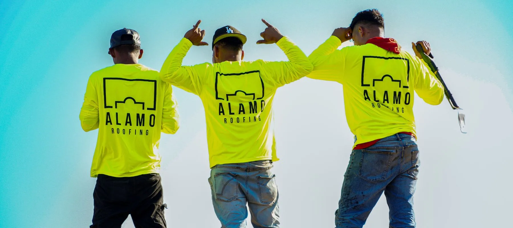 Three people stand with their backs facing the camera, wearing bright yellow Alamo Roofing shirts. Two point upward with both hands, and one holds a tool over their shoulder against a clear blue sky.
