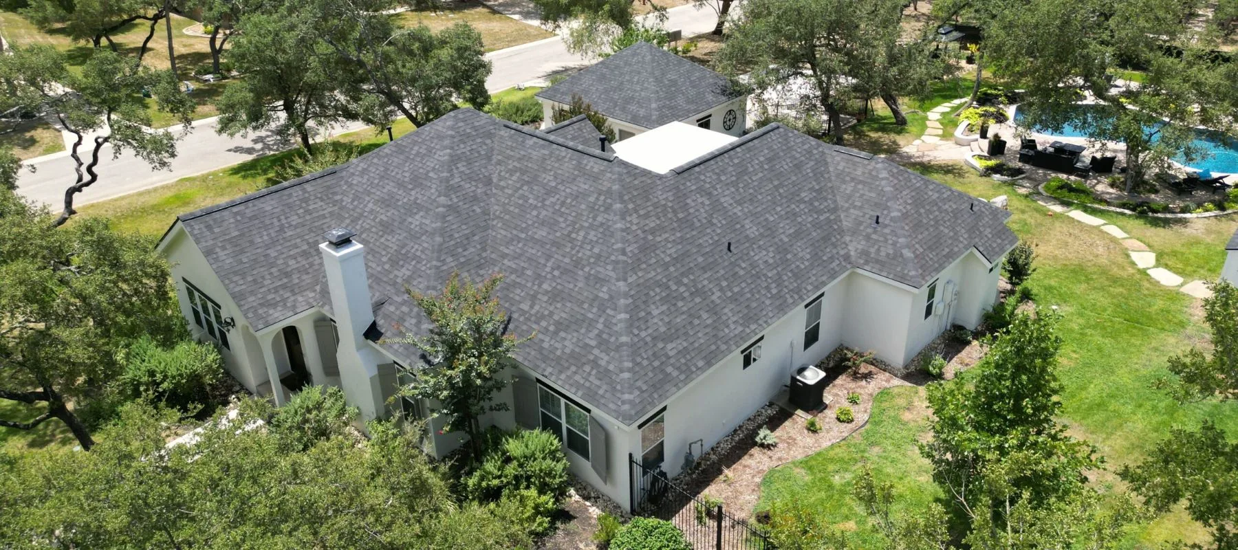 Aerial view of a large, white house with a dark gray shingle roof surrounded by trees, green lawn, and landscaping. There is a patio area and a pool visible in the background.