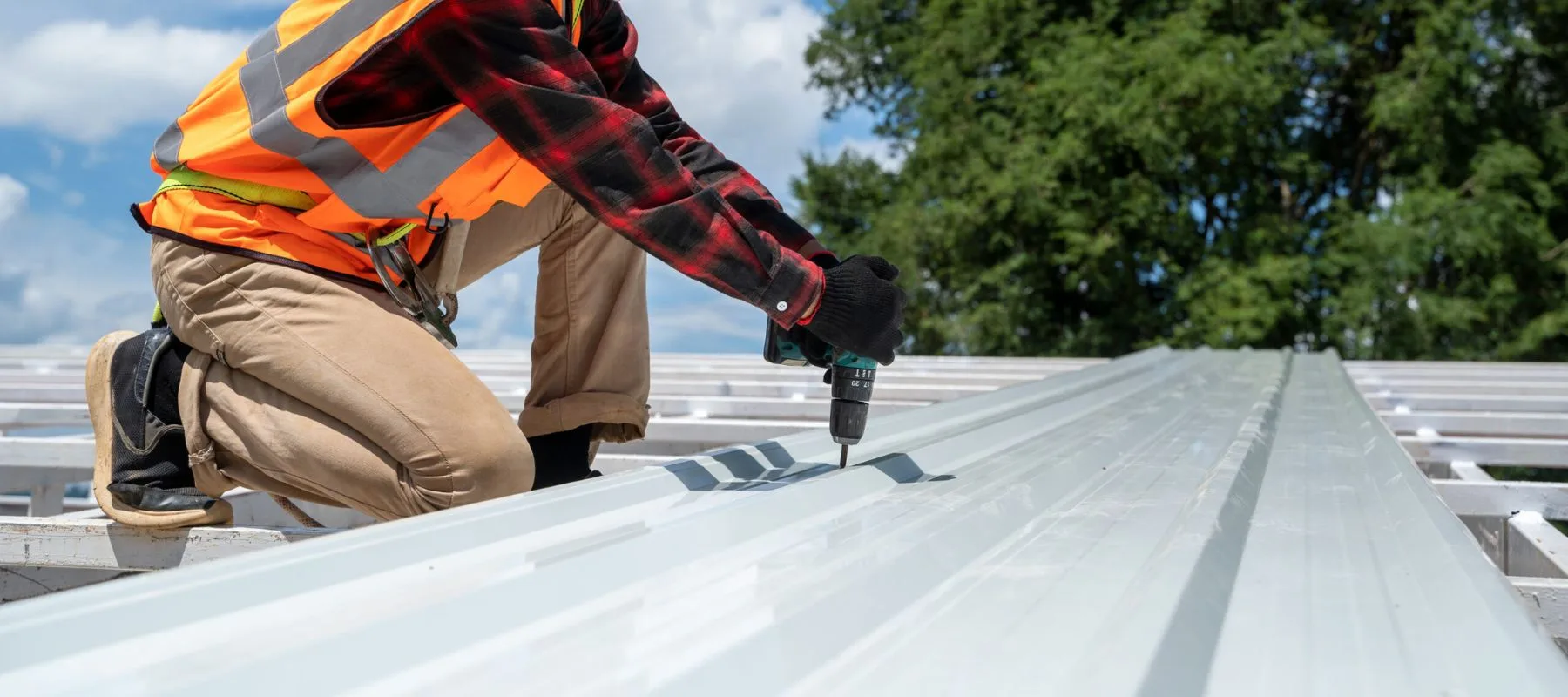A construction worker in a safety vest and gloves uses a power drill to fasten a metal sheet to a rooftop framework under a blue sky with trees in the background.