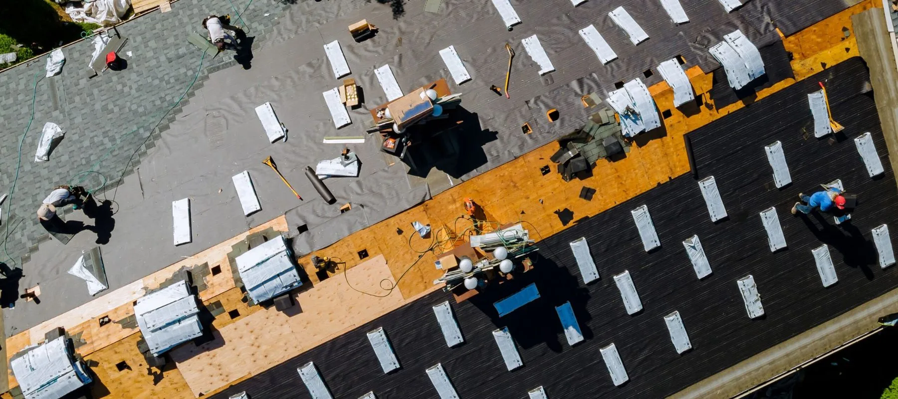 Aerial view of workers installing roofing materials on a large building; rolls of roofing, tools, and supplies are spread out on different colored roof sections.