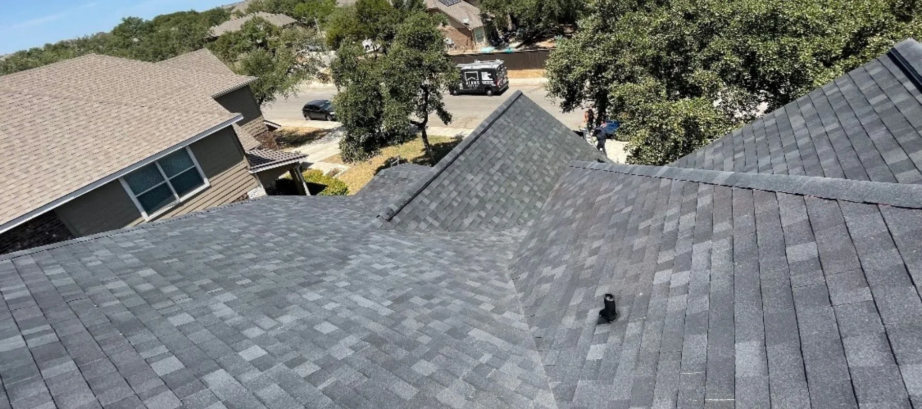 View from a rooftop with dark gray asphalt shingles, showing intersecting rooflines, nearby houses, trees, a street, and a parked vehicle in a residential neighborhood.
