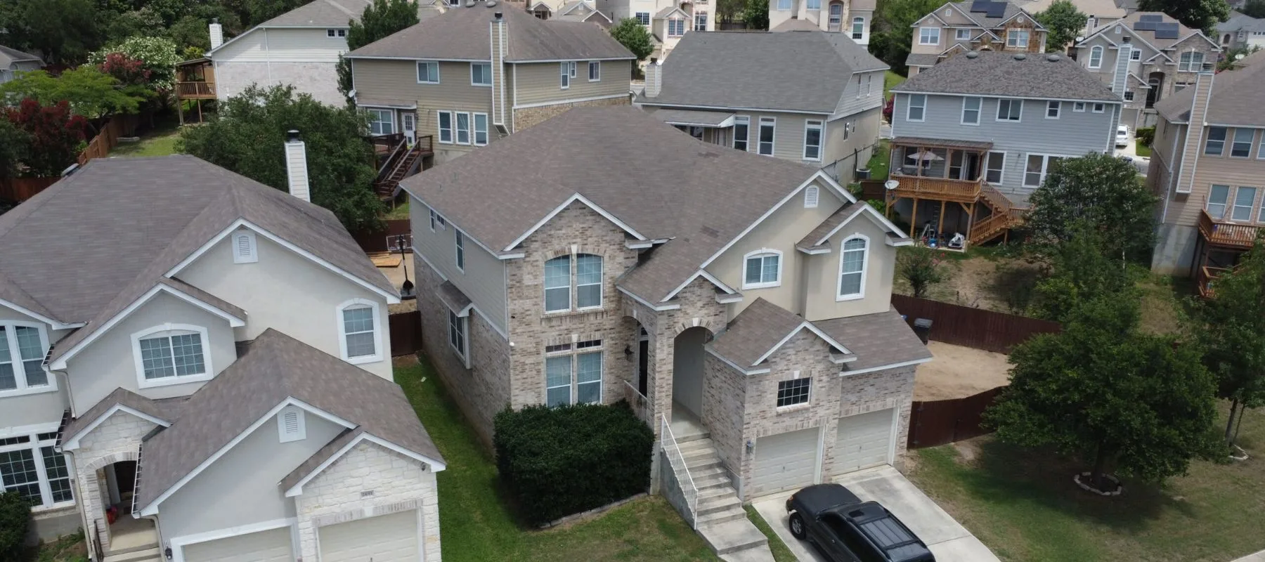 Aerial view of suburban two-story houses with beige and light brown exteriors, a black truck parked in a driveway, manicured lawns, and fenced backyards on a cloudy day.