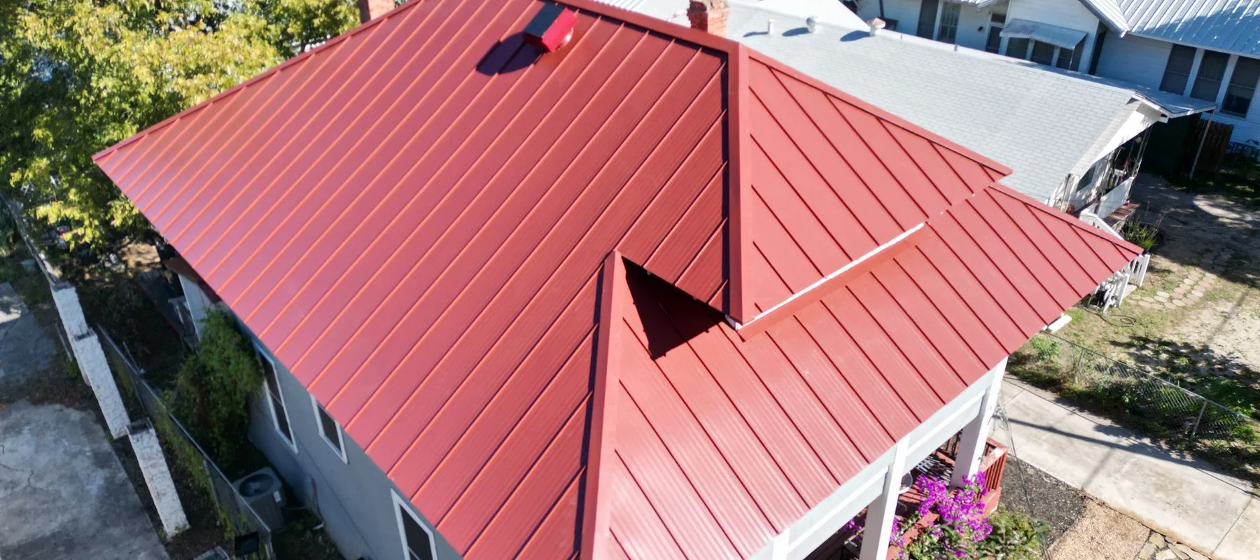 Aerial view of a house with a red metal roof featuring clean lines and multiple slopes, surrounded by trees, neighboring buildings, and some purple flowers in the yard.