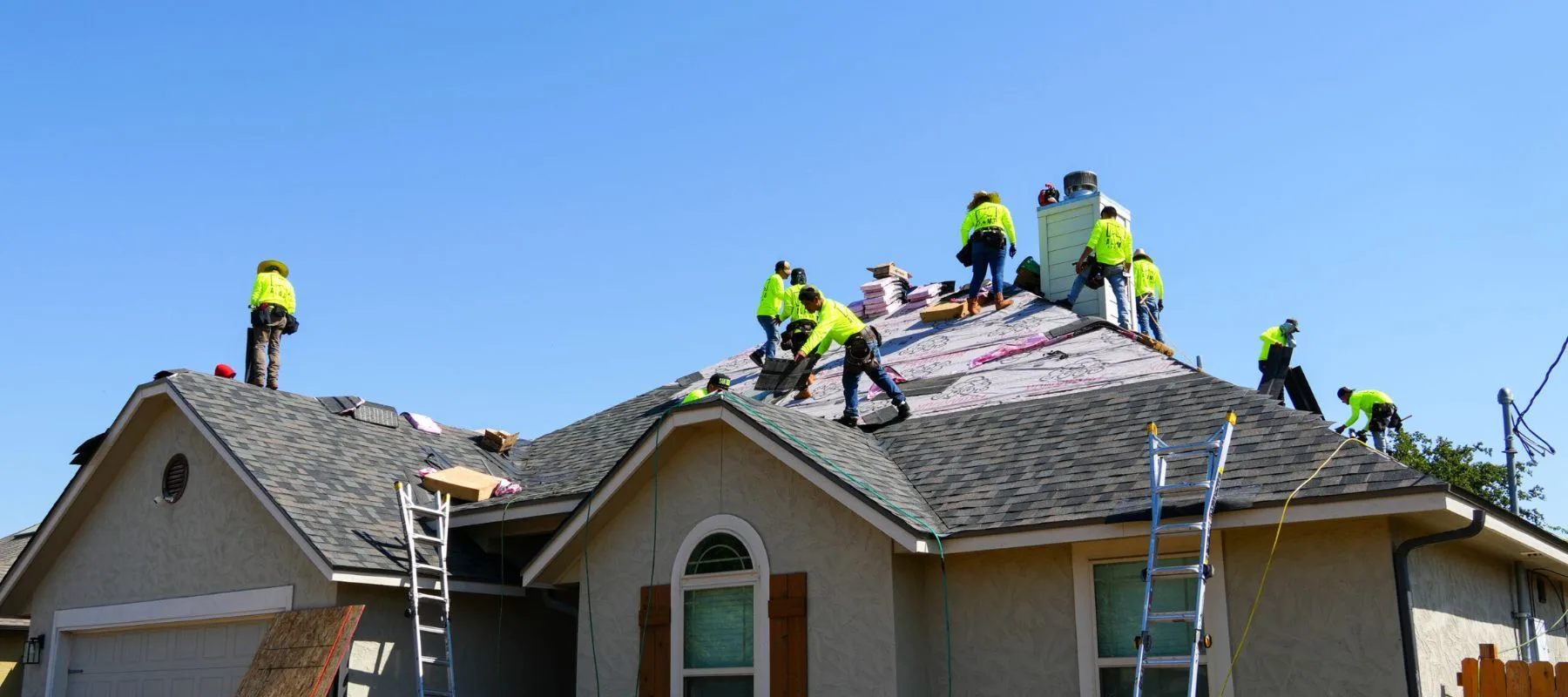 Several workers in bright yellow shirts repair the roof of a house under a clear blue sky, using ladders and roofing materials. The house has grey shingles and arched windows.