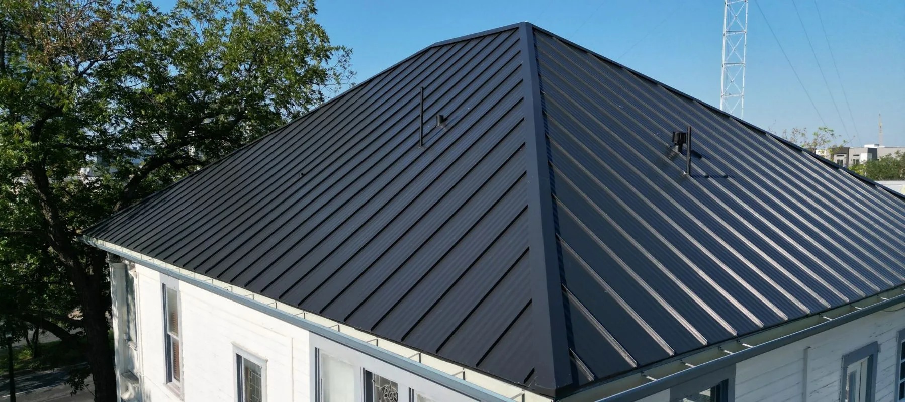 A white building with a dark gray standing seam metal roof, featuring clean lines and a pyramid-like shape, is shown under a clear blue sky with trees in the background.