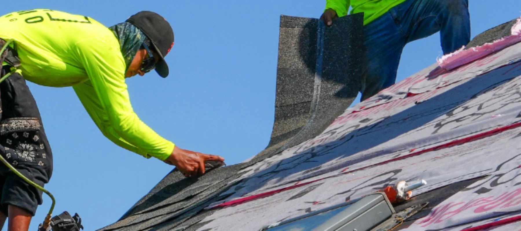 Two workers in neon shirts are installing asphalt shingles on a sloped roof under a clear blue sky. One worker is bent over securing a shingle while the other holds more shingles above.