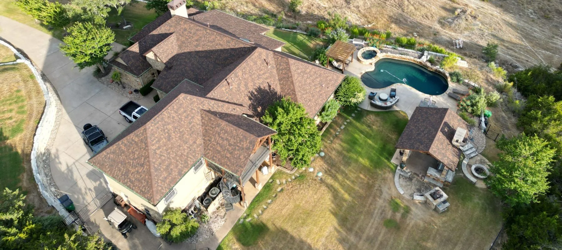 Aerial view of a large house with a brown roof, driveway with parked cars, green lawn, trees, backyard pool, spa, gazebo, and outdoor seating area, all surrounded by dry grassy land.