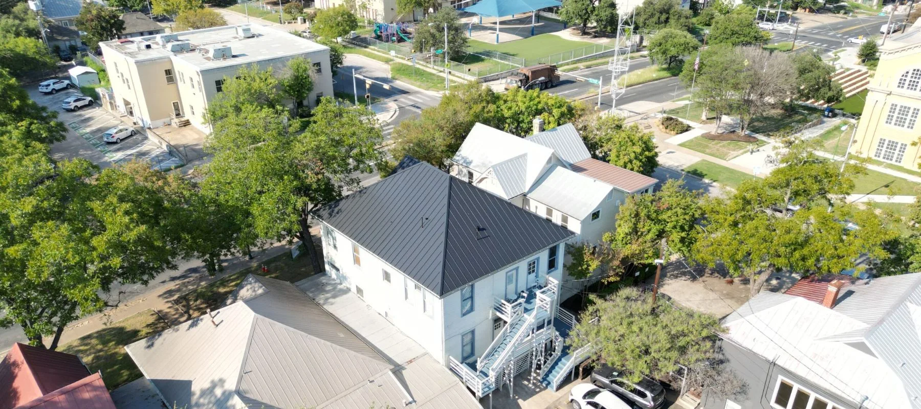 Aerial view of a residential area with white houses, tree-lined streets, parked cars, and a nearby park with a playground and sports courts. An outdoor stairway is visible on one building.