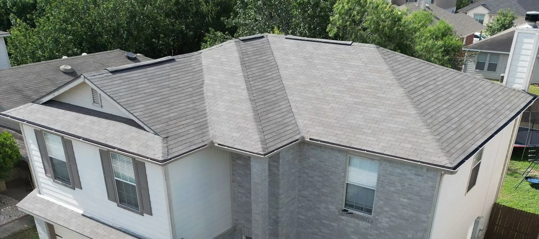 Aerial view of a house with a gray shingle roof, multiple slopes, and dormer sections, surrounded by trees and neighboring houses. The exterior walls are gray brick and white siding.