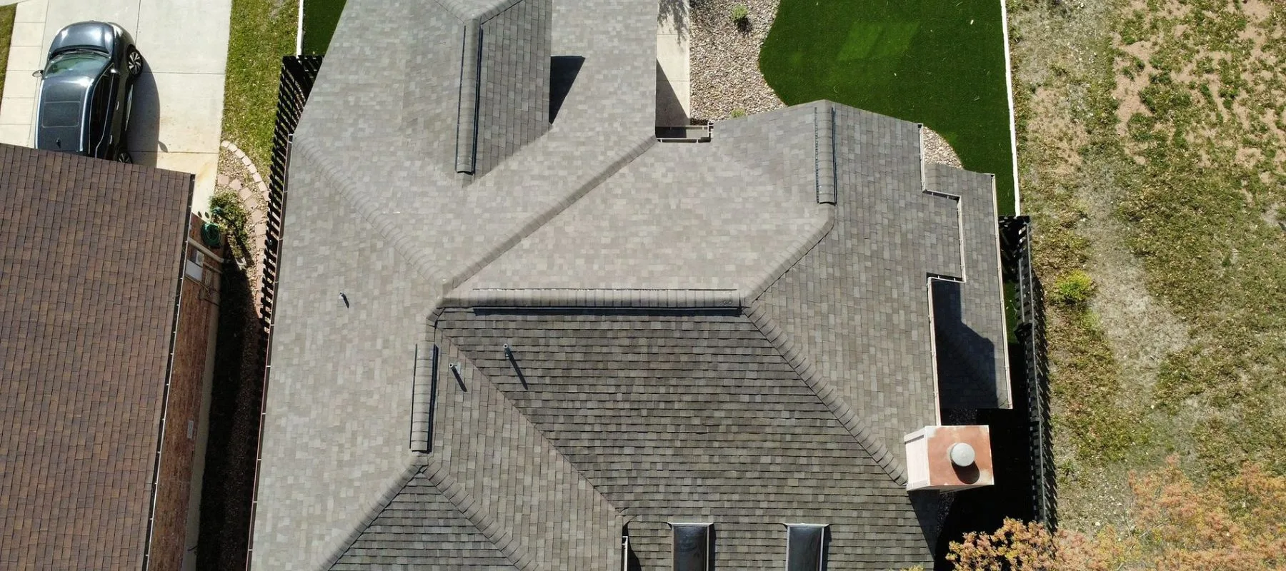 Aerial view of a house with a gray shingle roof, adjacent driveway and parked car, artificial green lawn on one side, and bare earth or dry grass area on the other side.