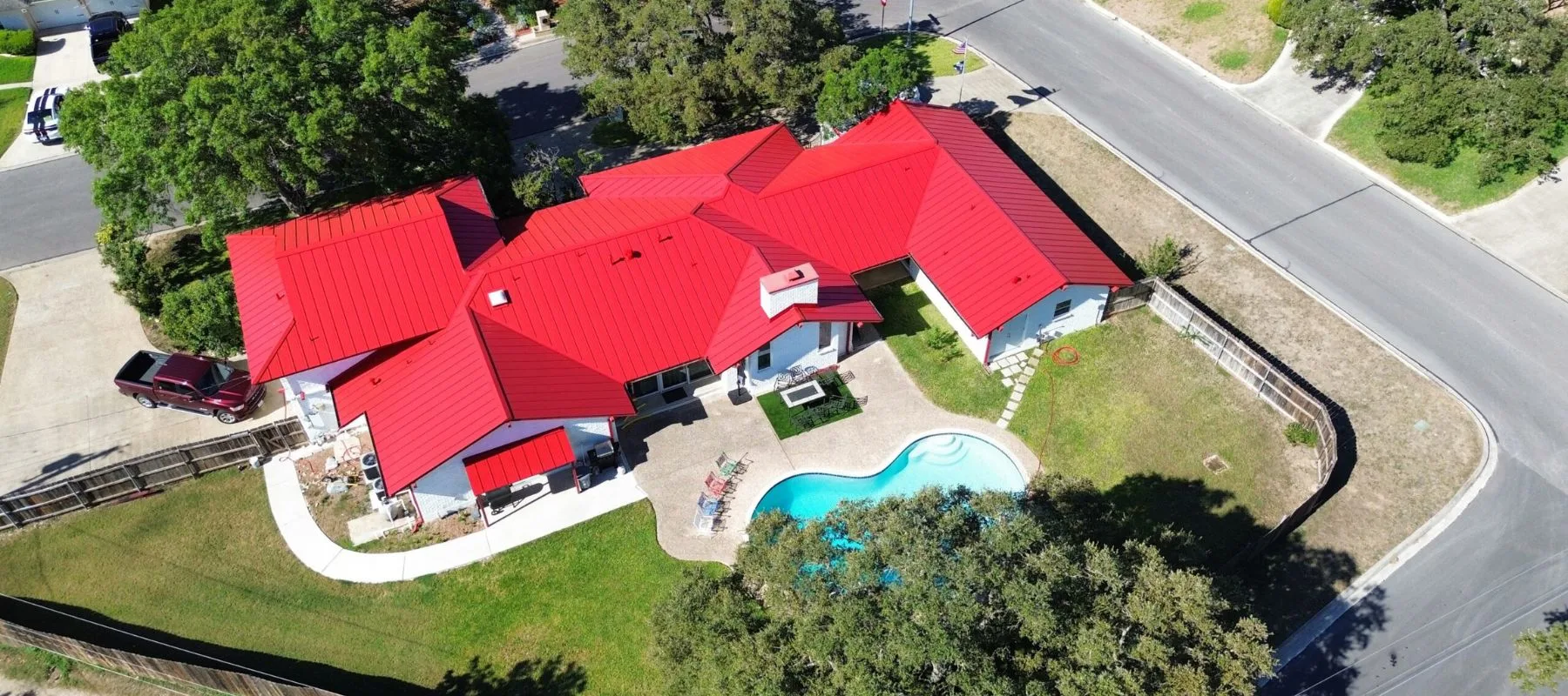 Aerial view of a large house with a bright red roof, white exterior walls, a backyard swimming pool, curved walkways, and trees surrounding the property, located at a street corner.