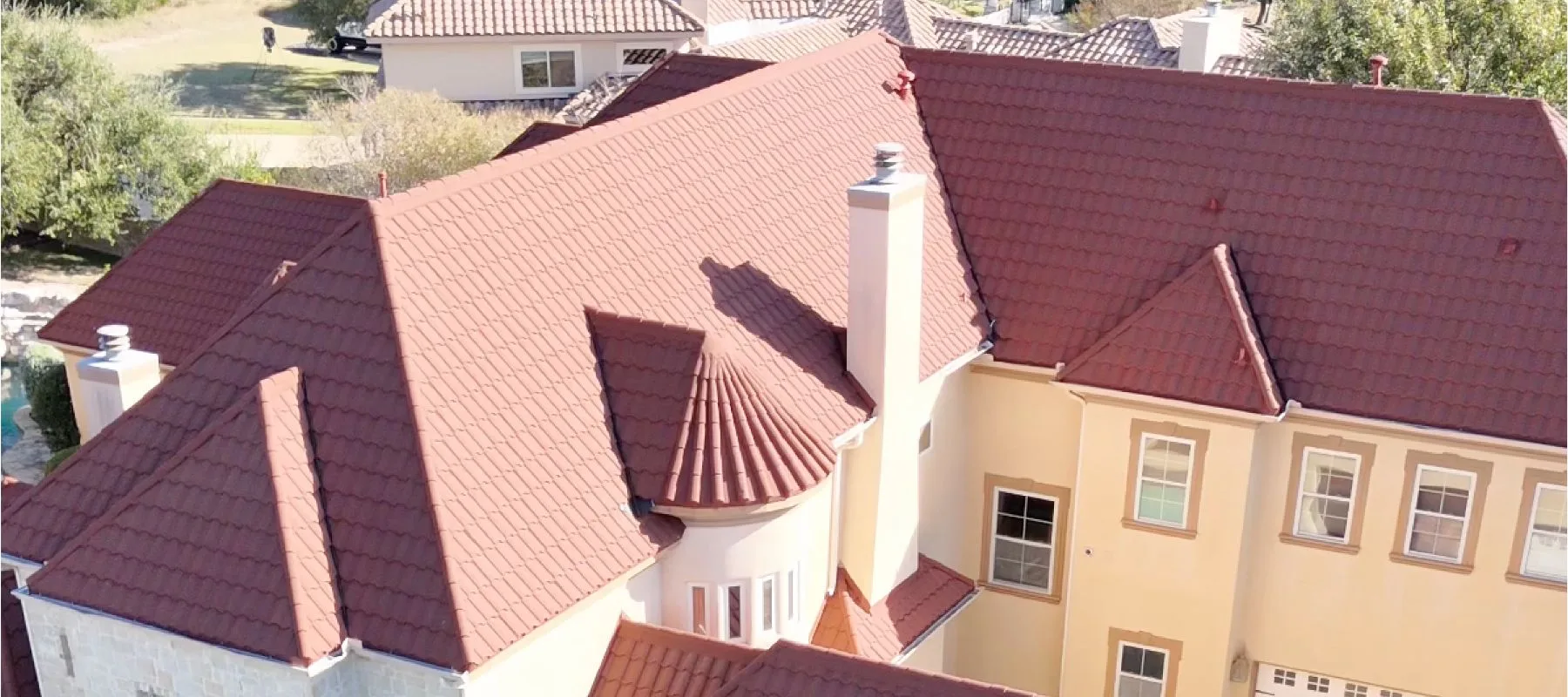 Aerial view of a large beige house with multiple red tiled roofs, chimneys, and a small turret, surrounded by trees and neighboring homes.