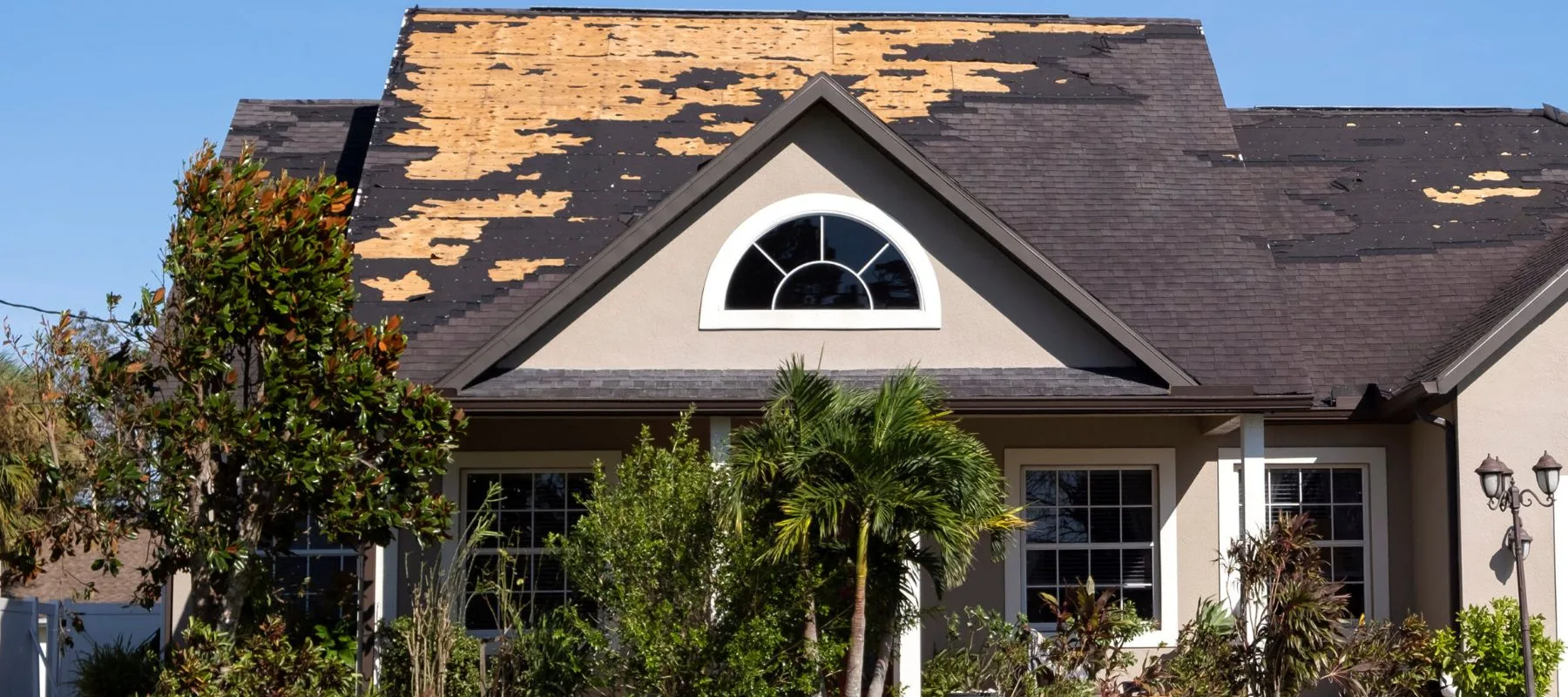 A house with significant roof damage; many shingles are missing from the upper section, exposing the wood underneath. The home is surrounded by green trees and shrubs under a clear blue sky.