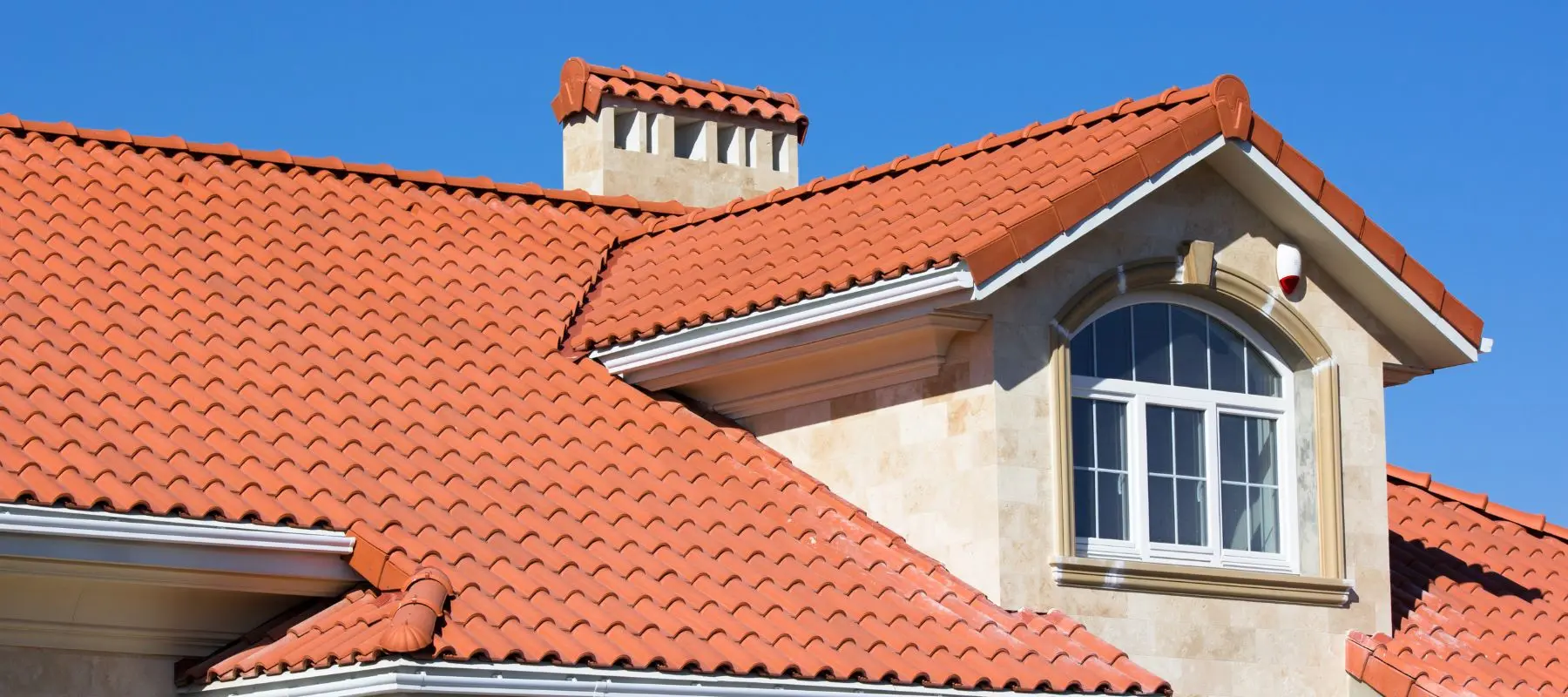 Close-up view of a house roof with red clay tiles, light-colored stone walls, a chimney, and an arched window under a clear blue sky.