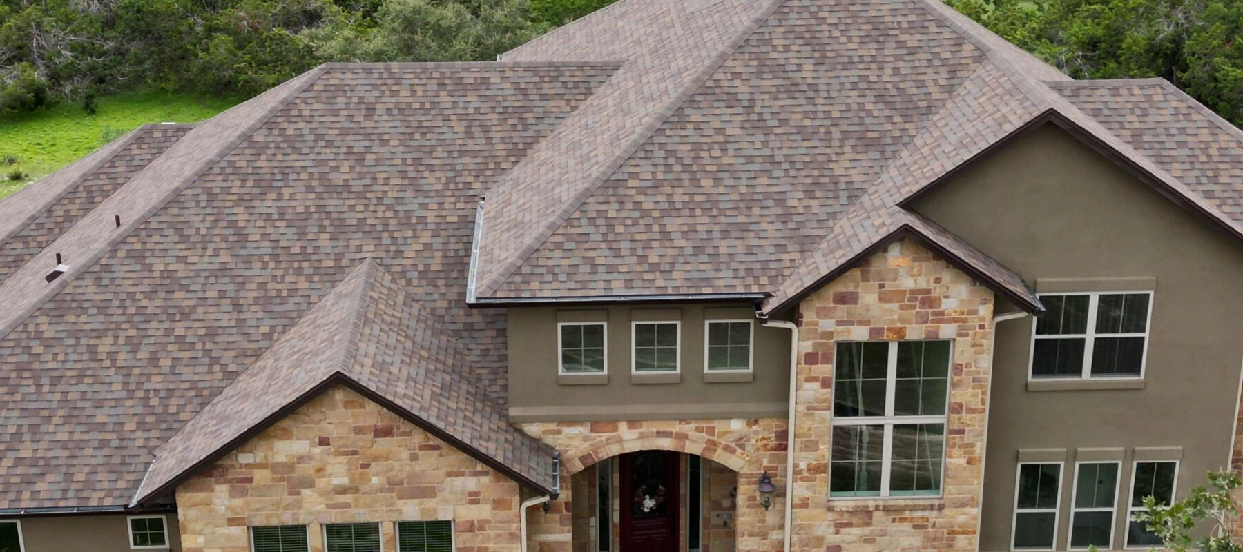 A large two-story house with a complex brown shingle roof, stone and stucco exterior, large windows, and an arched entryway surrounded by greenery.