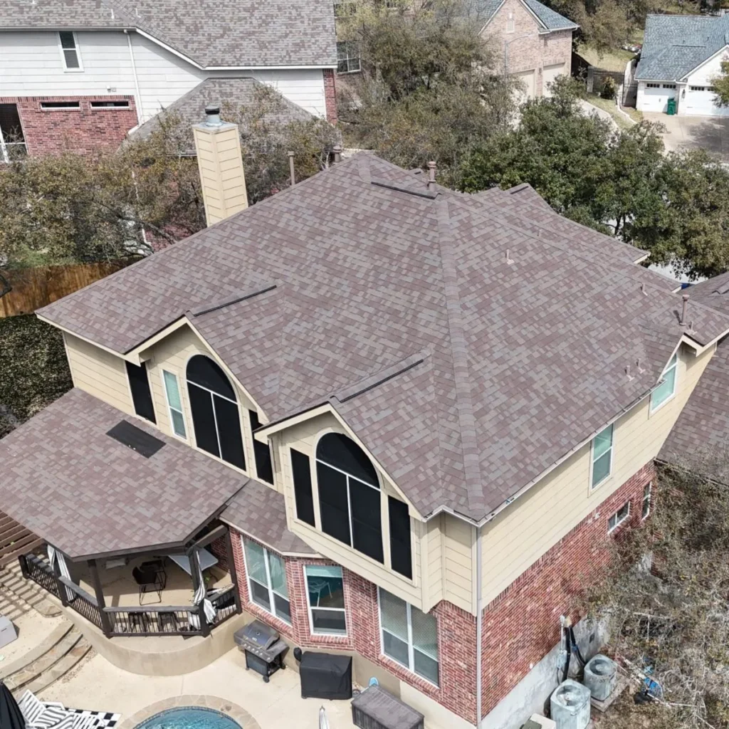 Aerial view of a two-story house with a brown shingle roof, tan siding, red brick lower exterior, large windows, a patio area, a round swimming pool, and nearby trees and neighboring houses.
